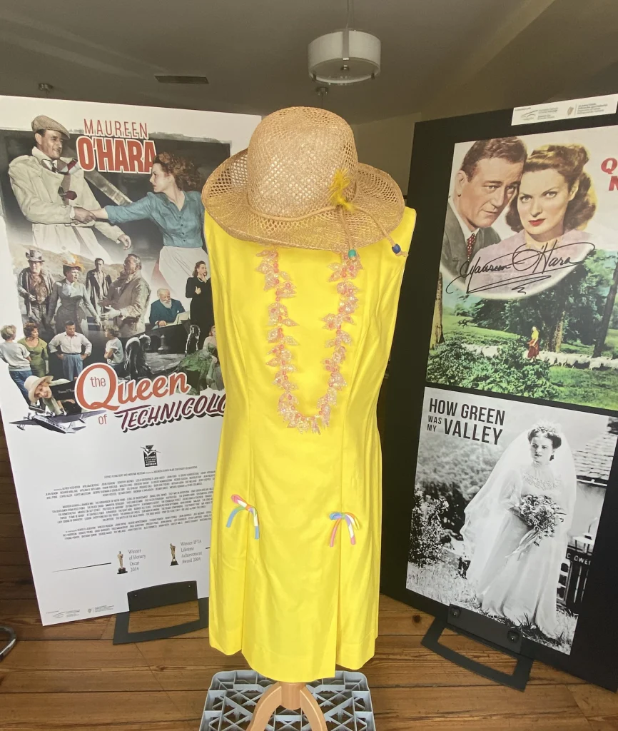 Full-length mannequin display of a bright yellow dress with an open back and a straw hat, in front of The Quiet Man posters.