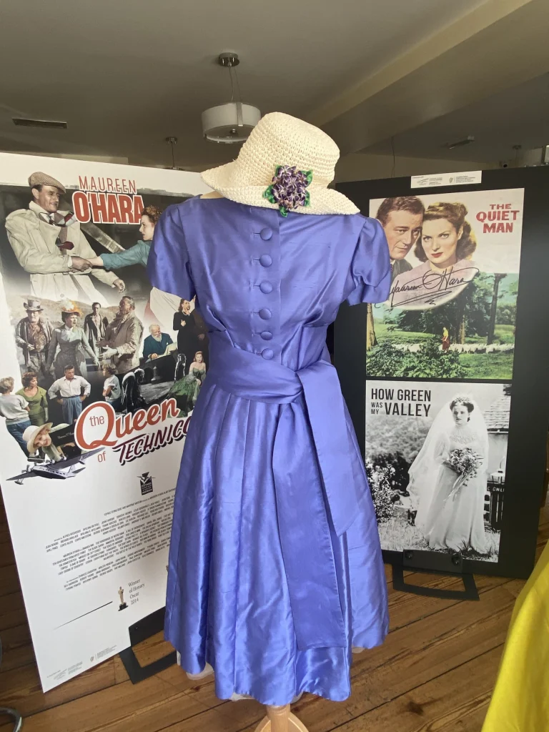 Full-length mannequin display of a royal purple satin gown with a wide belt and a white hat, placed in front of The Quiet Man movie posters.