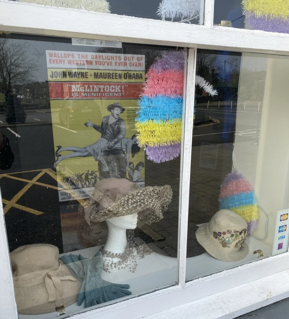 Shop window display featuring colorful knitted items, antique bonnets, and a vintage film poster for a musical.